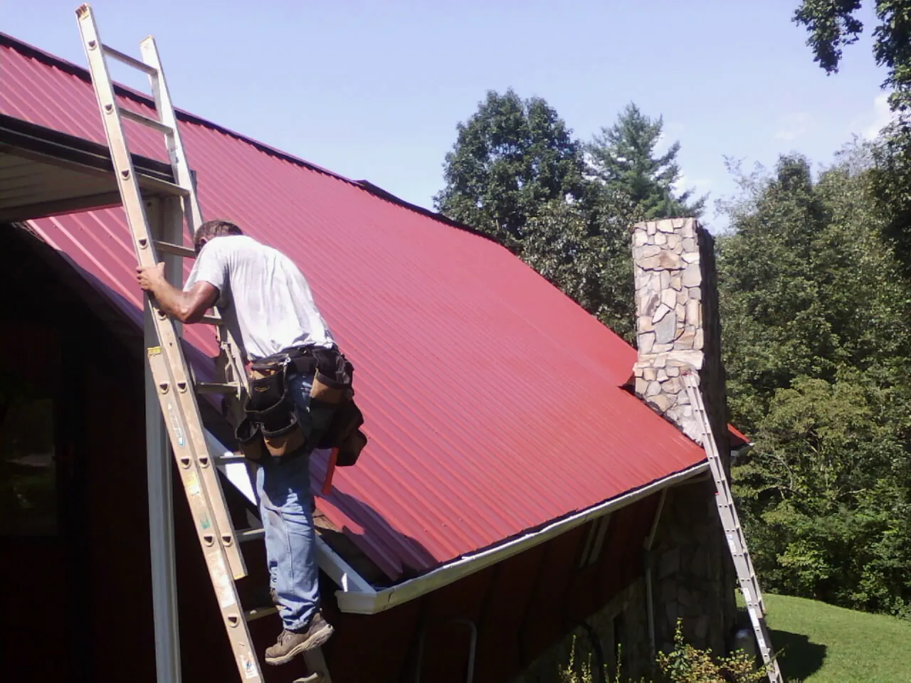 Roofer on a ladder beside a red metal roof and stone chimney — Soffit & Fascia Repair by Roofing Brothers in Watkins